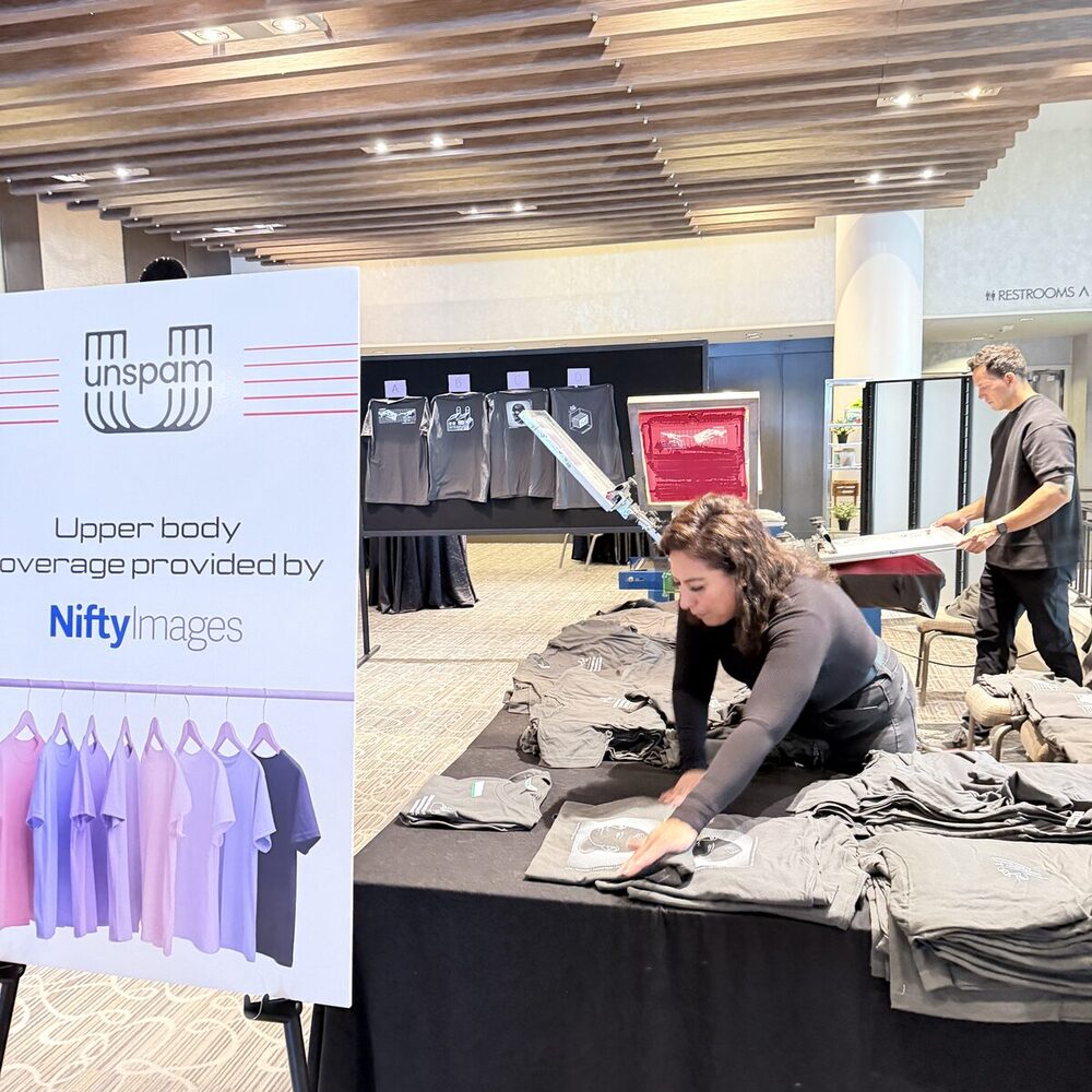 A Merch Troop crew member folds fresh-printed event tees at a draped table while a press runs in the background