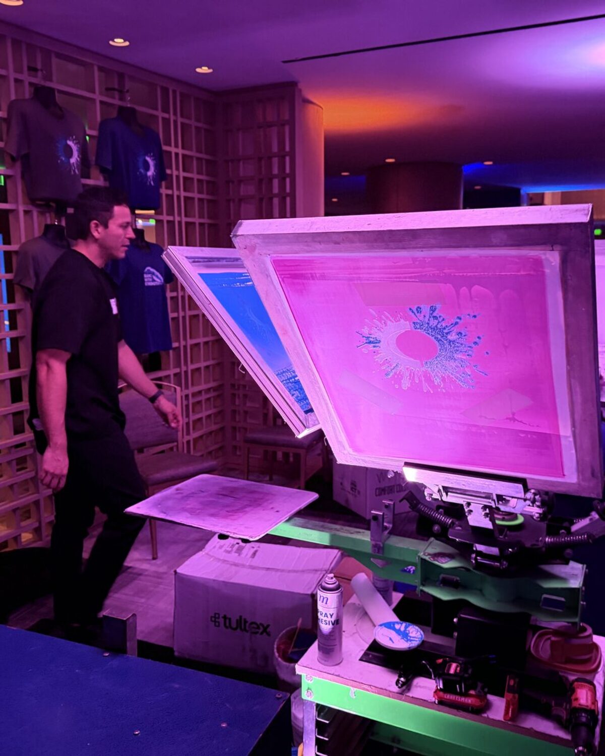A Merch Troop printer at a live screen-printing press lit in pink and purple, with freshly printed tees displayed behind the station