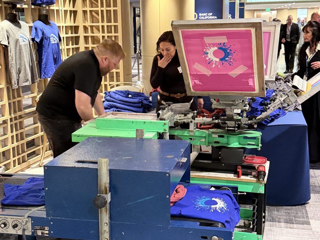 Two Merch Troop printers running a screen press with a pink screen, finished blue tees displayed behind them