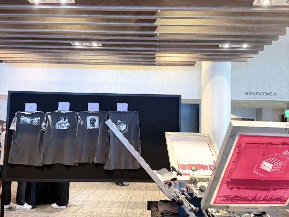 Printed sample tees displayed on a board beside a screen press under a warm wood-slat ceiling